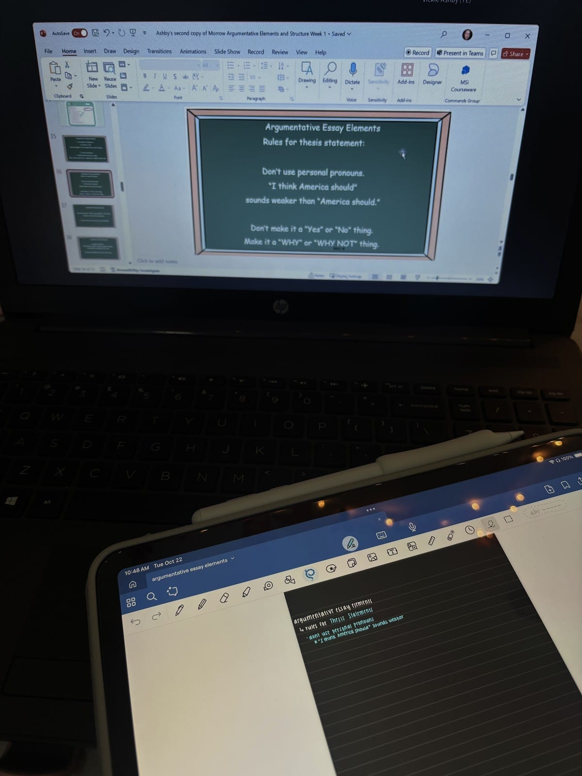 A laptop and tablet showing notes for an argumentative essay. The laptop screen has a PowerPoint slide titled "Argumentative Essay Elements," with rules for thesis statements. The tablet displays handwritten notes on the same topic.