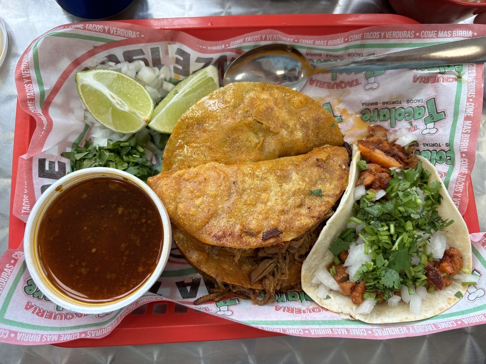 A tray with Mexican food, including two birria tacos with a side of broth, chopped onions, cilantro, lime wedges, and a small taco. The dish is garnished with fresh herbs, and a spoon is placed beside the food.