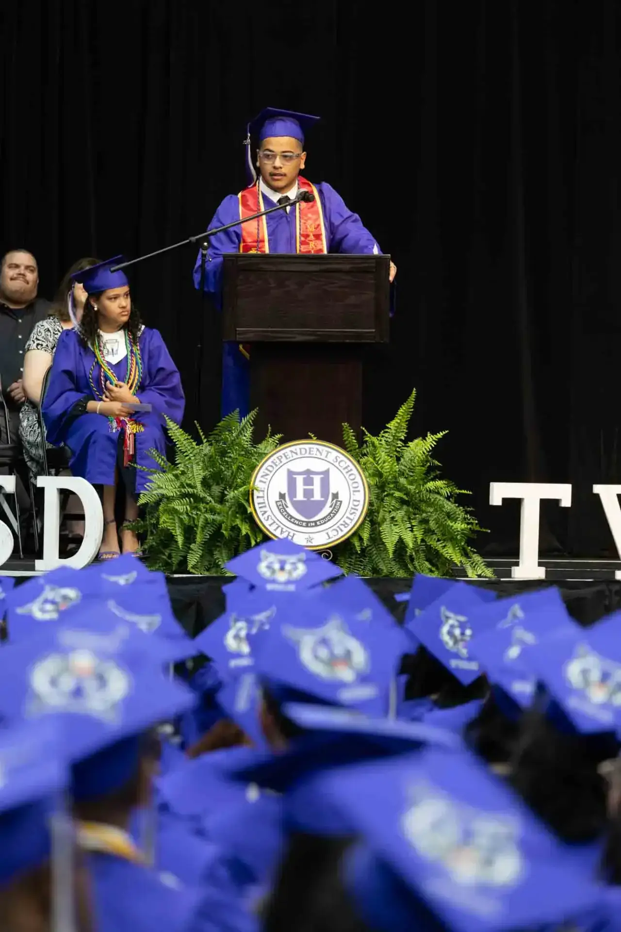 A graduate in a blue cap and gown speaks at a podium during a graduation ceremony. Seated graduates and a school emblem are visible, conveying celebration.