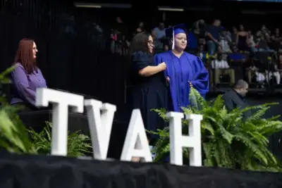 Graduate in blue cap and gown receives congratulations on stage amid lush ferns and large "TVAH" letters, watched by an audience in a formal ceremony.