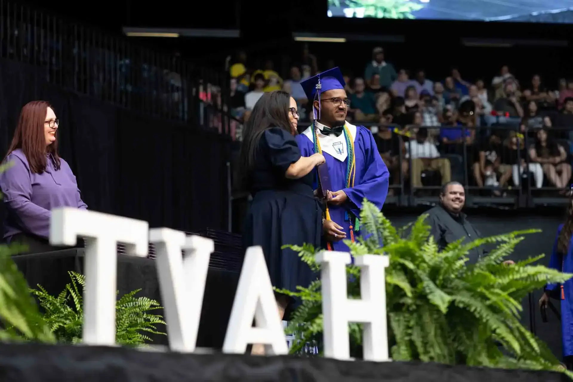 Graduate in cap and gown receives a diploma on stage at a ceremony, smiling with faculty. "TVAH" letters and green ferns adorn the foreground.