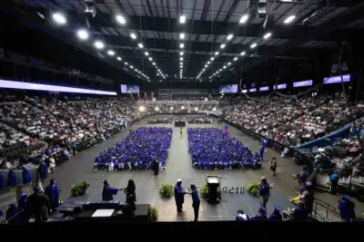 Wide-angle view of a graduation ceremony in an indoor arena. Graduates in blue caps and gowns are seated in the center, surrounded by a large audience. The setting is formal and celebratory.
