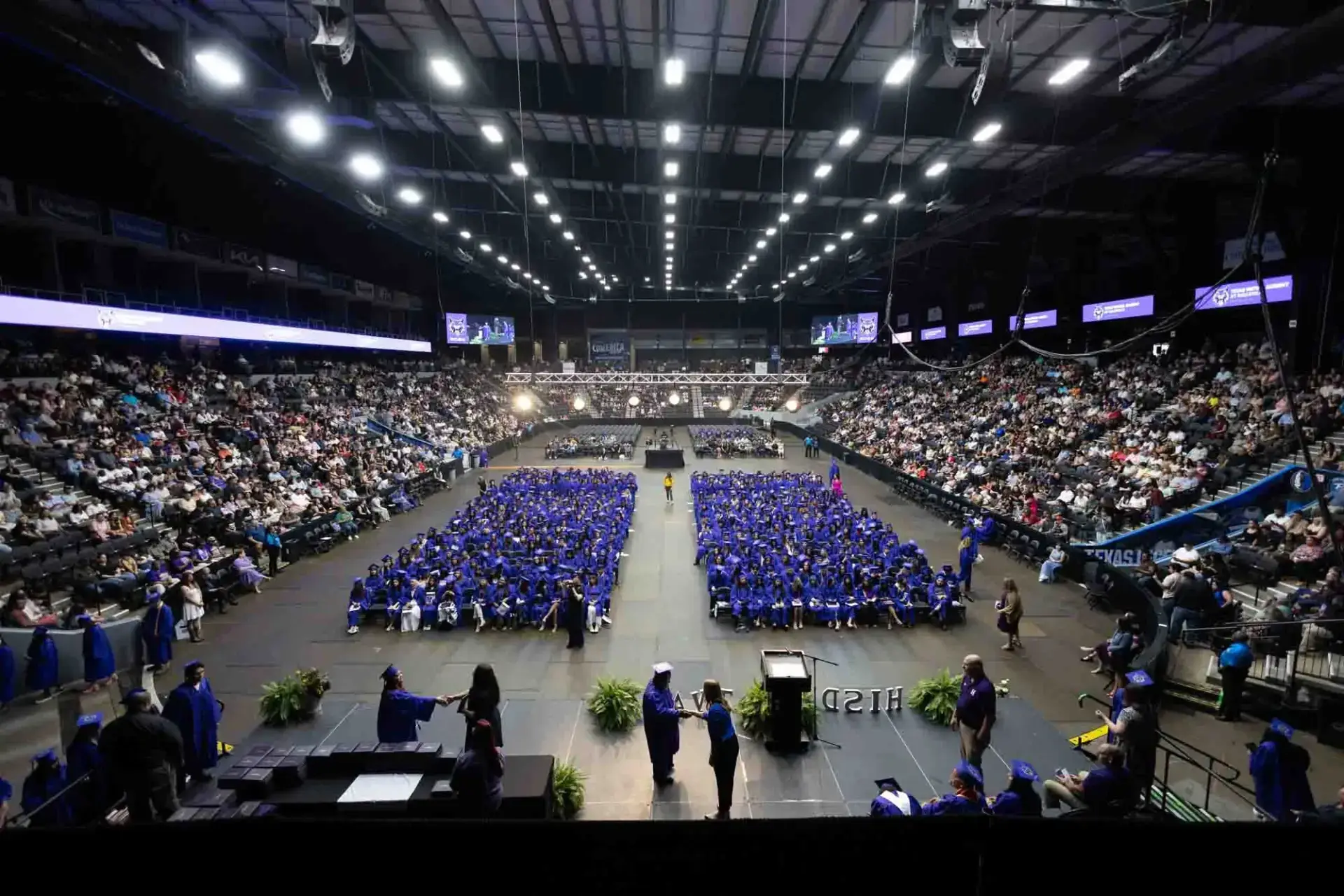 Wide-angle view of a graduation ceremony in an indoor arena. Graduates in blue caps and gowns are seated in the center, surrounded by a large audience. The setting is formal and celebratory.