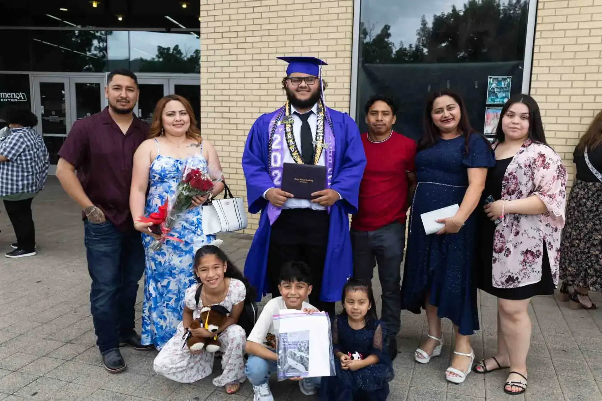 A graduate in a blue cap and gown holds a diploma, surrounded by seven smiling family members. Some hold flowers and a teddy bear, celebrating outside.