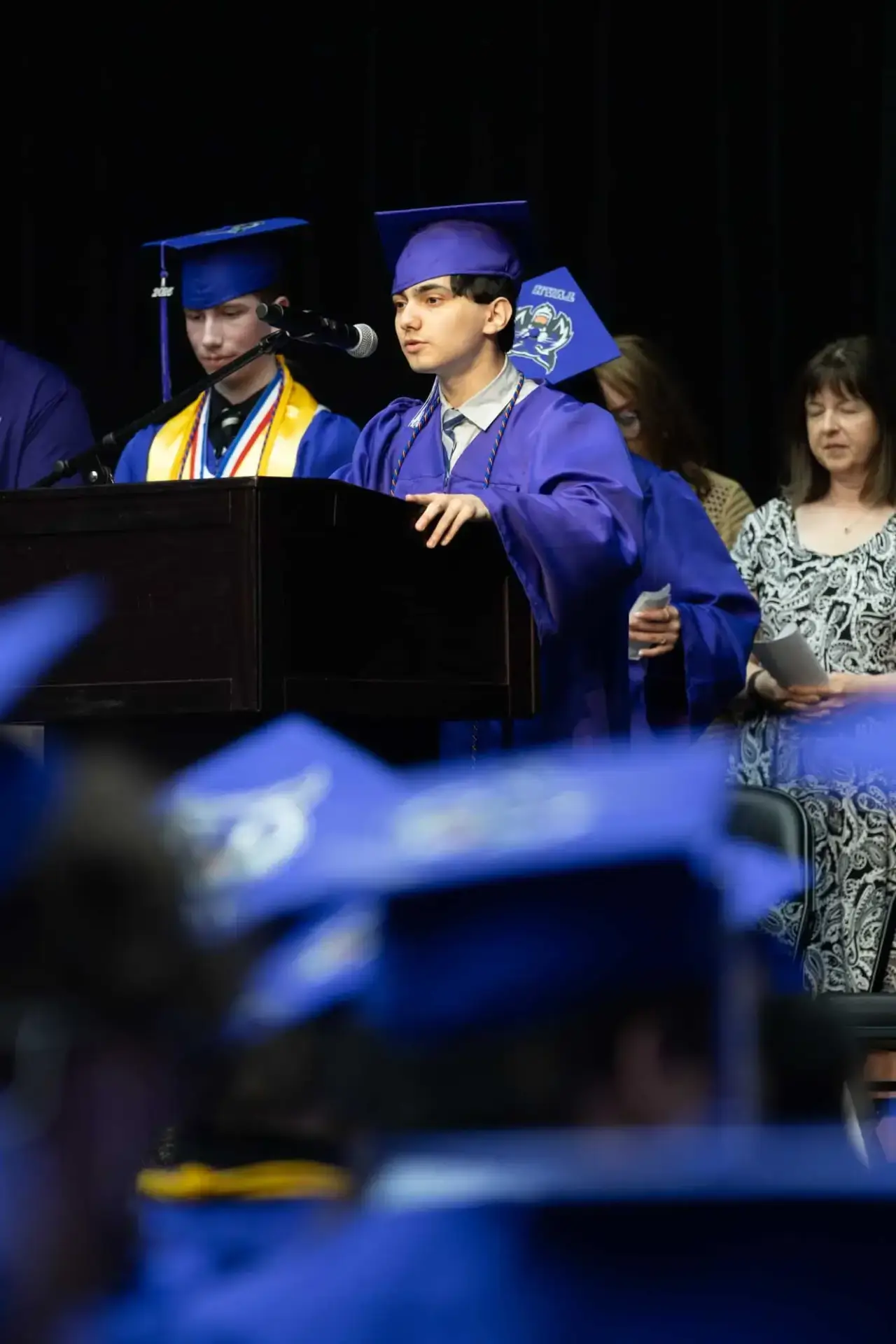 A graduate in blue cap and gown speaks at a podium on stage during a ceremony. A peer stands beside them, and people watch in the background.