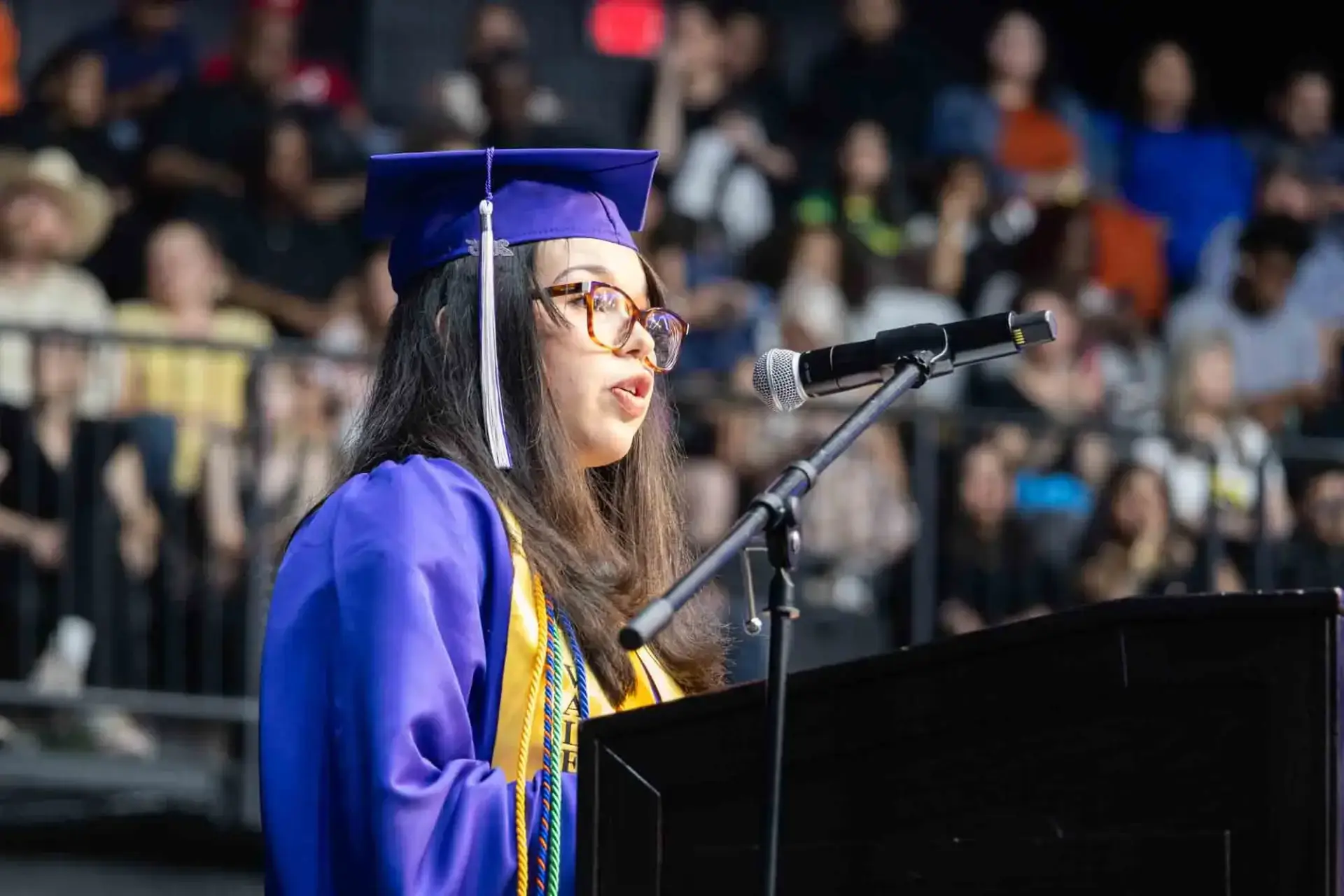 A graduate in a blue cap and gown speaks confidently at a podium, addressing the audience. The background shows a blurred crowd in attendance.