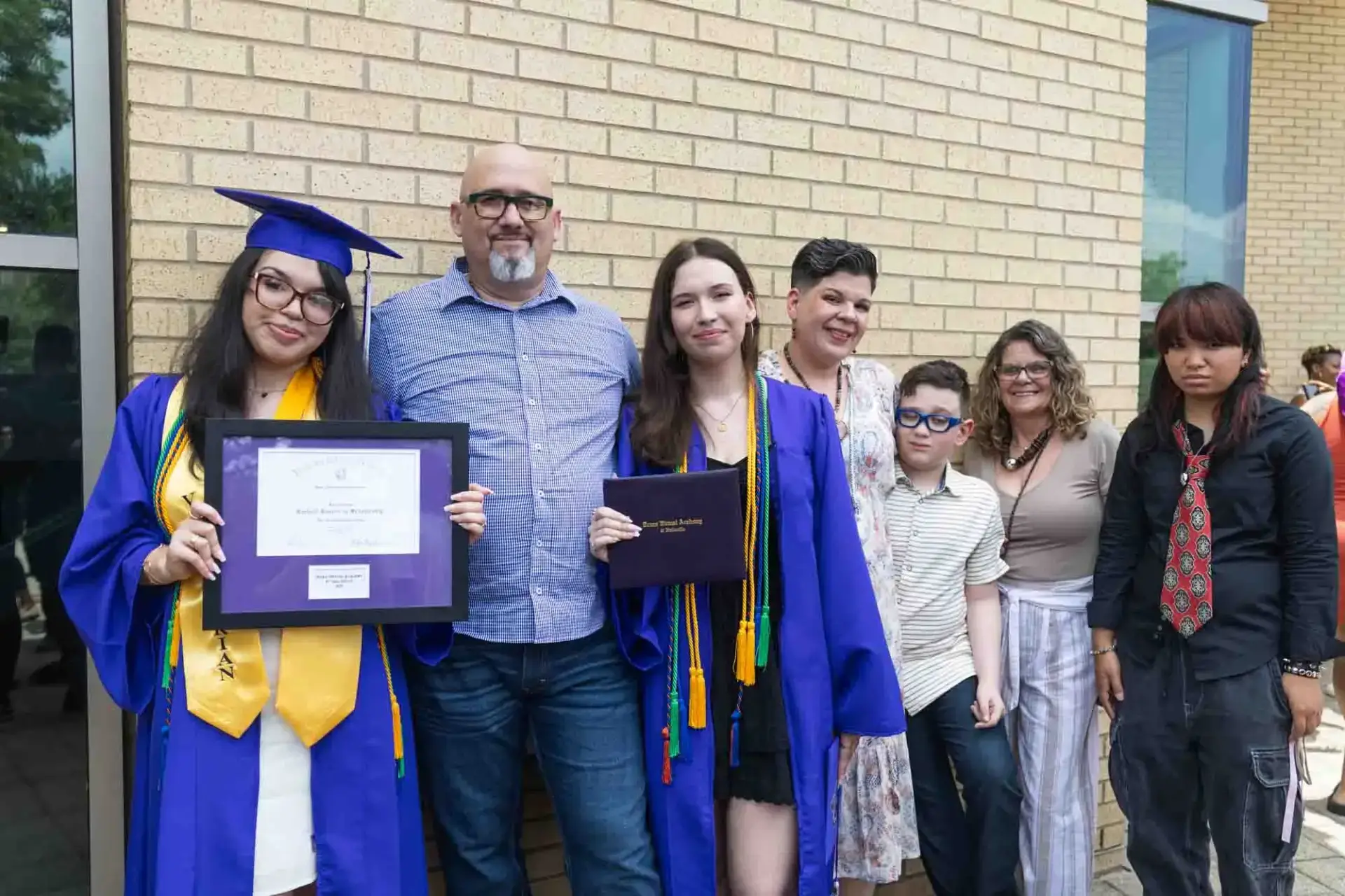Two graduates in blue caps and gowns hold diplomas, smiling proudly. They're surrounded by supportive family members against a brick wall backdrop.