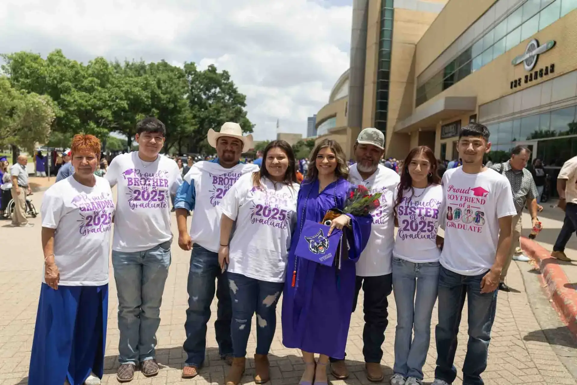 A group of eight people pose outdoors. A woman in a purple graduation gown holds flowers, surrounded by supportive family members wearing custom t-shirts. The atmosphere is celebratory.