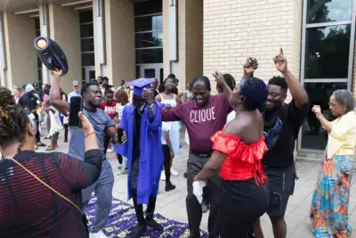 Graduation celebration outside a building. A graduate in a blue cap and gown is joyfully surrounded by cheering friends and family, capturing a lively moment.